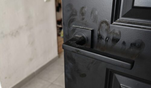 Detailný záber na znečistené čierne interiérové dvere s matnou čiernou kľučkou, kde sú na povrchu okolo rozety a na samotnej kľučke výrazne viditeľné mastné odtlačky prstov, dlane a jemná vrstva usadeného prachu.